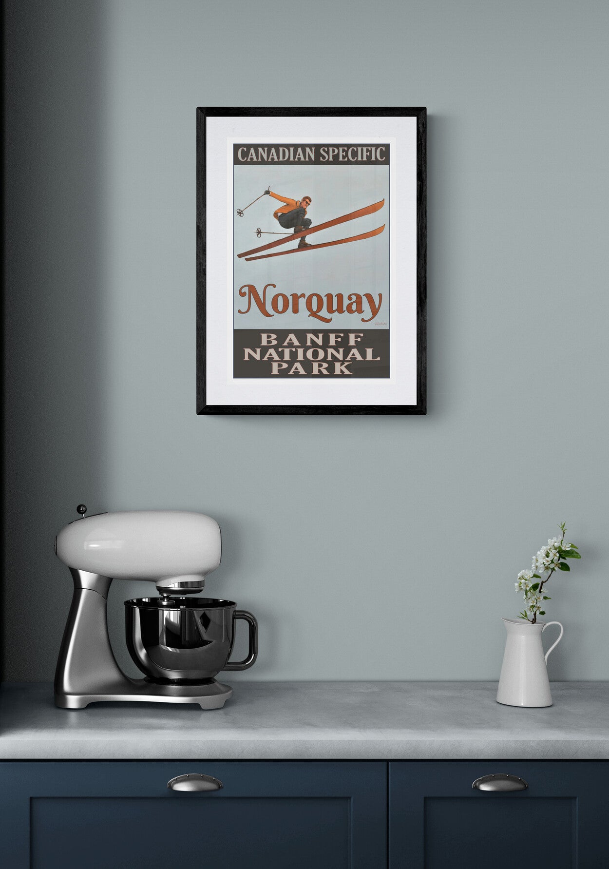 Framed poster of a skier in Norquay, Banff National Park on a gray wall above a kitchen counter with a mixer and vase.